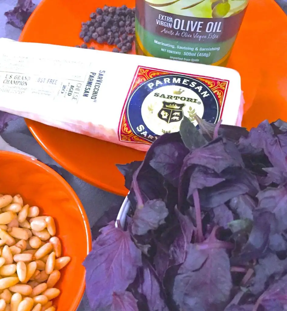 Ingredients for purple basil pesto: Large purple leaves of basil, an orange bowl with cream coloured pine nuts, an orange plate with a wedge of parmesan cheese, black peppercorns and part of a bottle of extra virgin olive oil with a green and black label