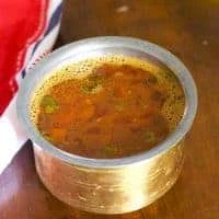 Brass vessel filled with reddish brown Tomato rasam. Pieces of tomato floating on top. A red and white napkin to the left, all of it on a brown wooden background