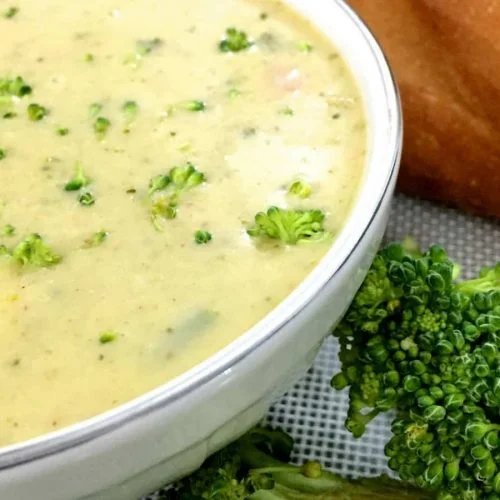 Yellow Broccoli Cheddar Soup flecked with green bits of herbs and broccoli,in a white bowl with a baguette in the background and pieces of green broccoli on the side