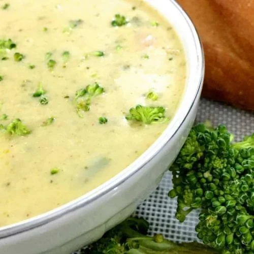 Yellow Broccoli Cheddar Soup flecked with green bits of herbs and broccoli,in a white bowl with a baguette in the background and pieces of green broccoli on the side