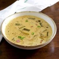Pale yellow creamy raw mango curry with okra, shallots, mango and curry leaves floating on top, in a white bowl edged with beige. On a dark brown background with a white napkin with green stripe behind